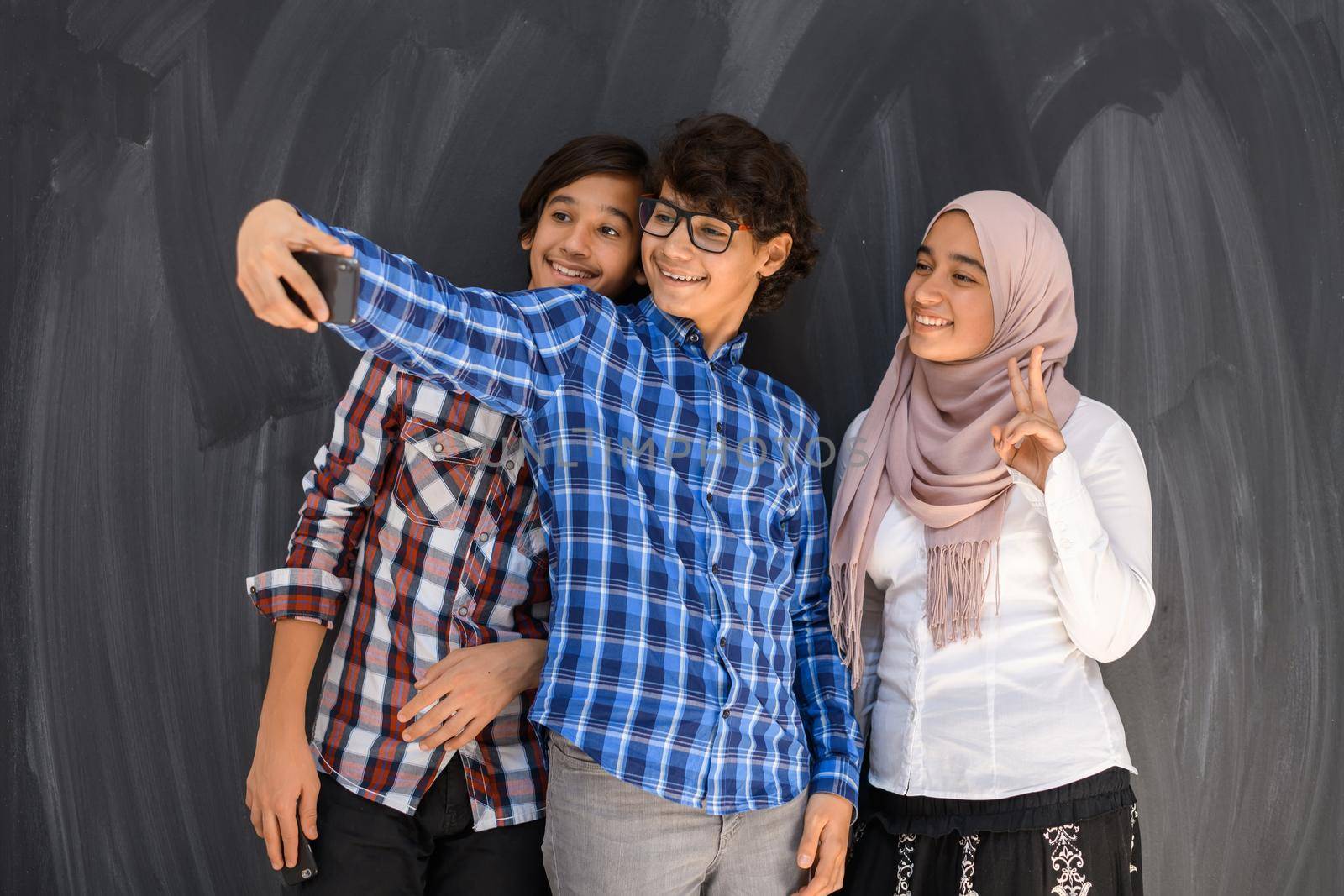 Group of Arab teens taking selfie photos on a smartphone with a black chalkboard in the background. Selective focus. High quality photo