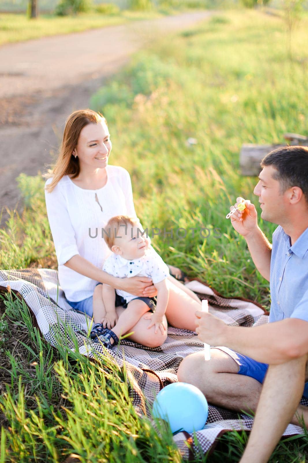 Young caucasian mother and father sittling on grass with little baby and blowing bubbles. Concept of picnic and children, parenthood and leisure time.