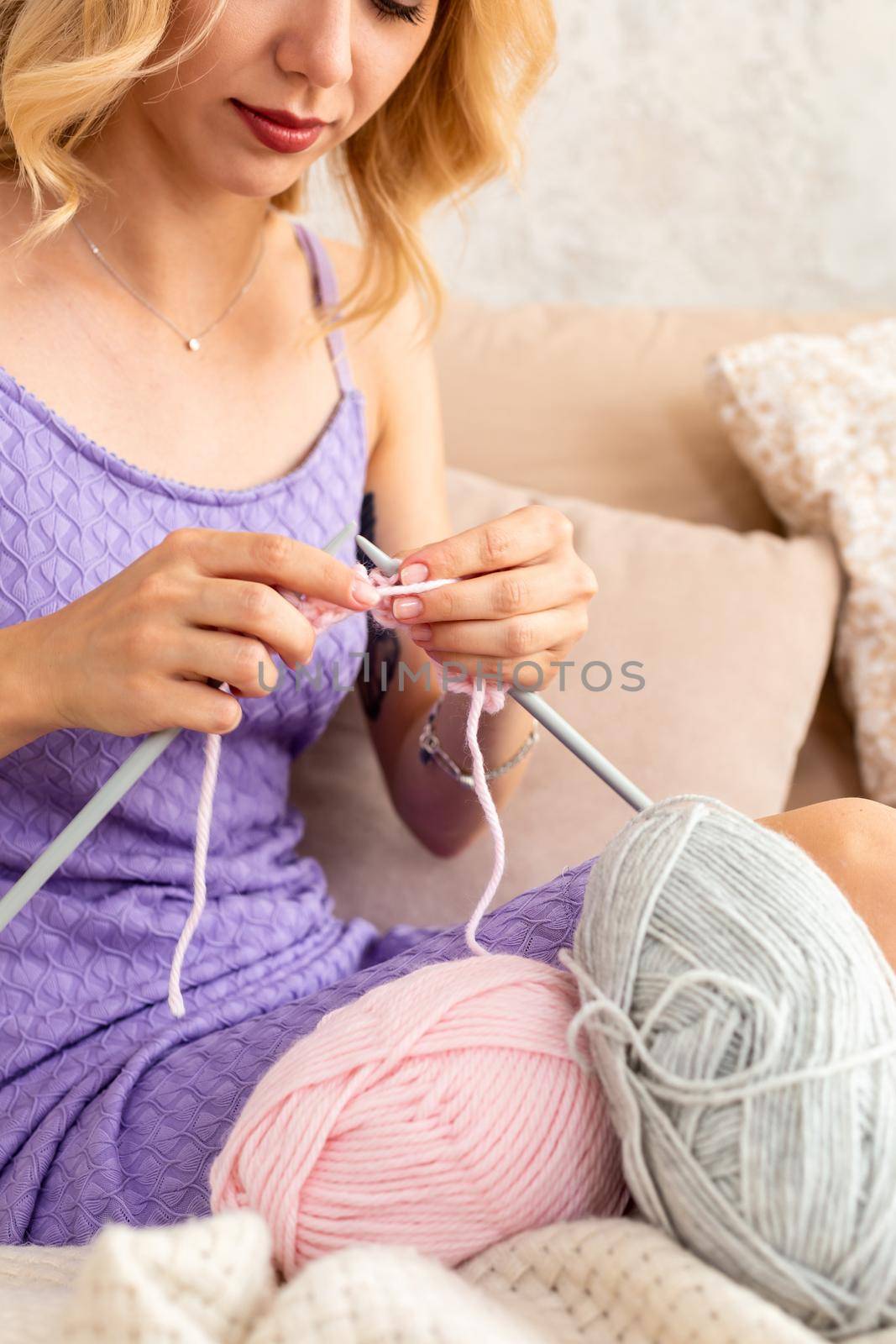 Close-up of woman in a lilac home dress knits a light pink scarf or plaid from natural threads on the bed. Close-up horizontal photo. Freelance creative working and living concept.