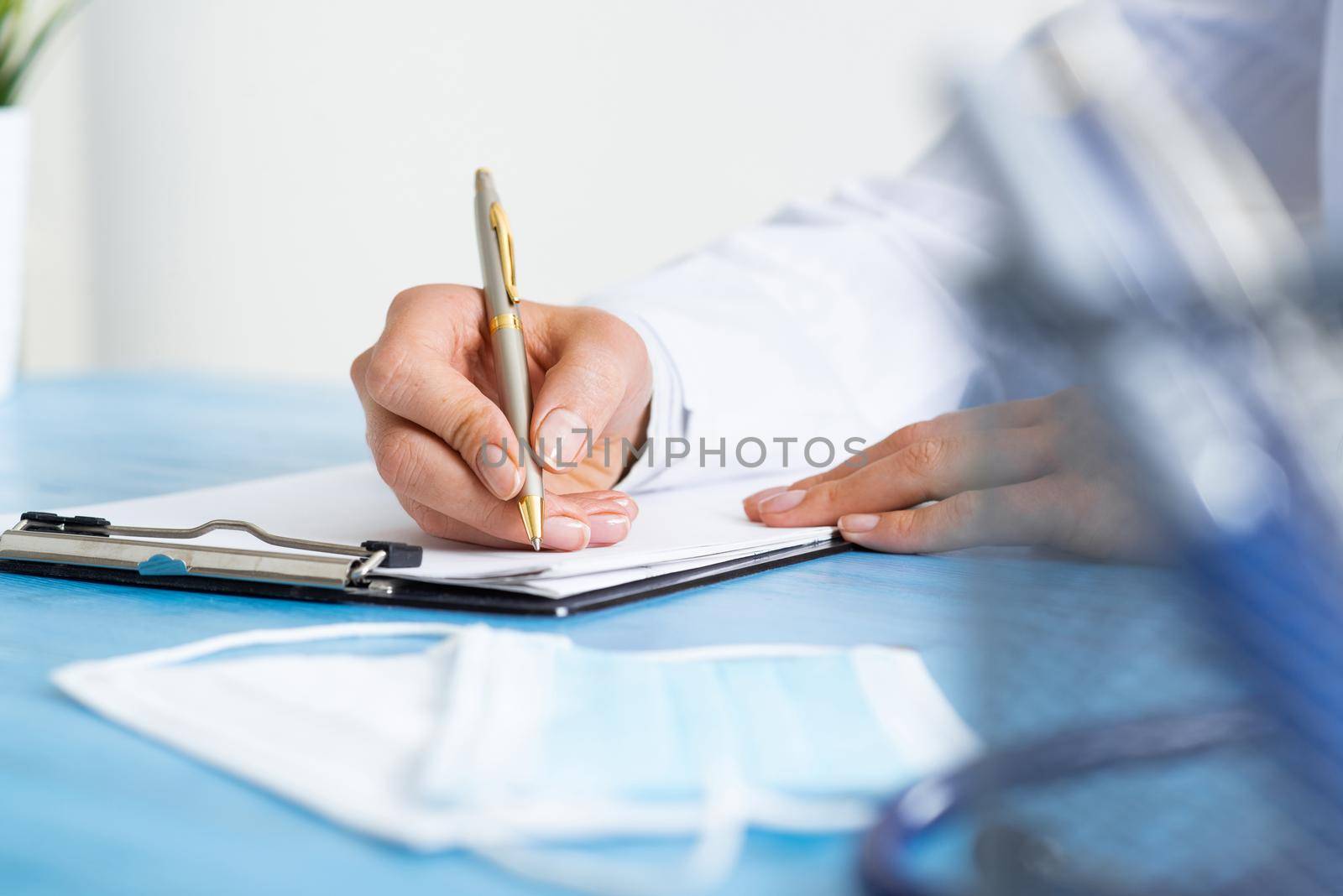 Close-up of female doctor filling medical form at clipboard. Therapist working at desk in office. Examination and diagnosis in modern clinic. Doctor workplace with green plant in pot and stethoscope.