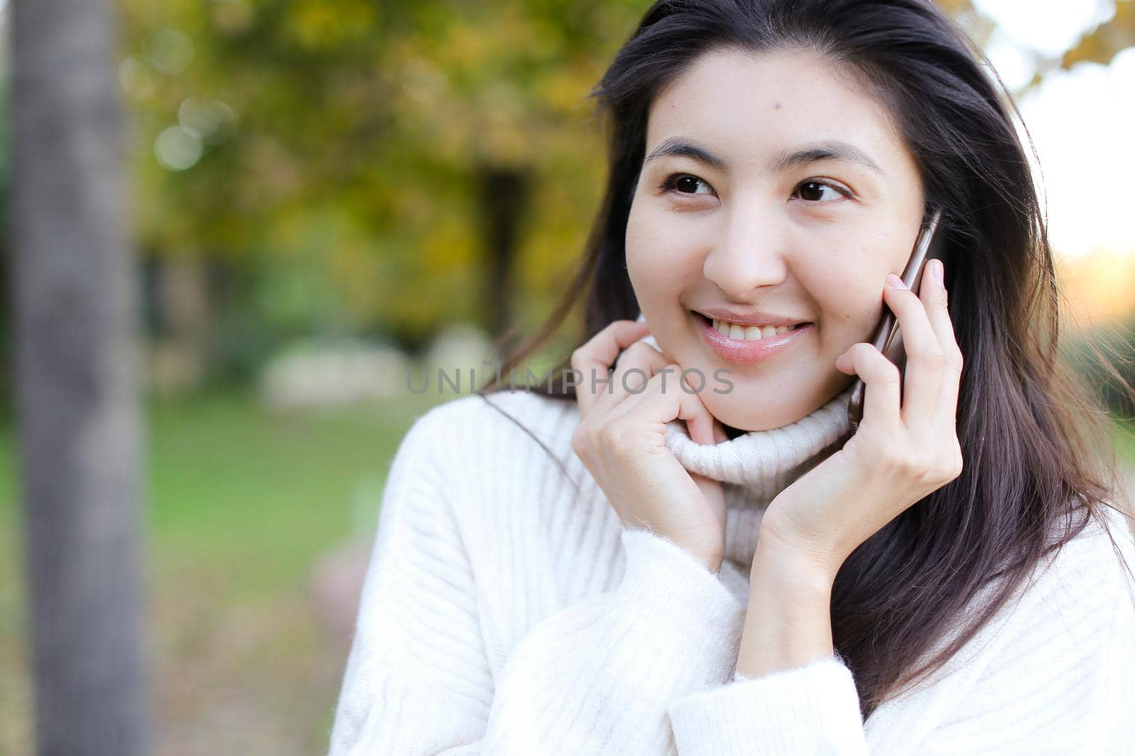 Close up asian smling woman wearing white sweater speaking by smartphone outside. Concept of chinese girl and modern technology.