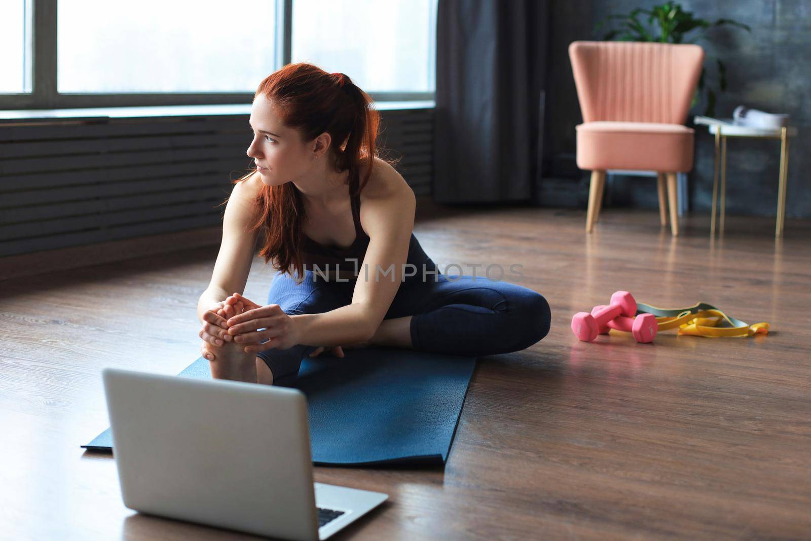 Beautiful slim sporty woman is stretching while sitting on the floor in the living room, watching in laptop where trainer showing exersices in zoom. Healthy lifestyle