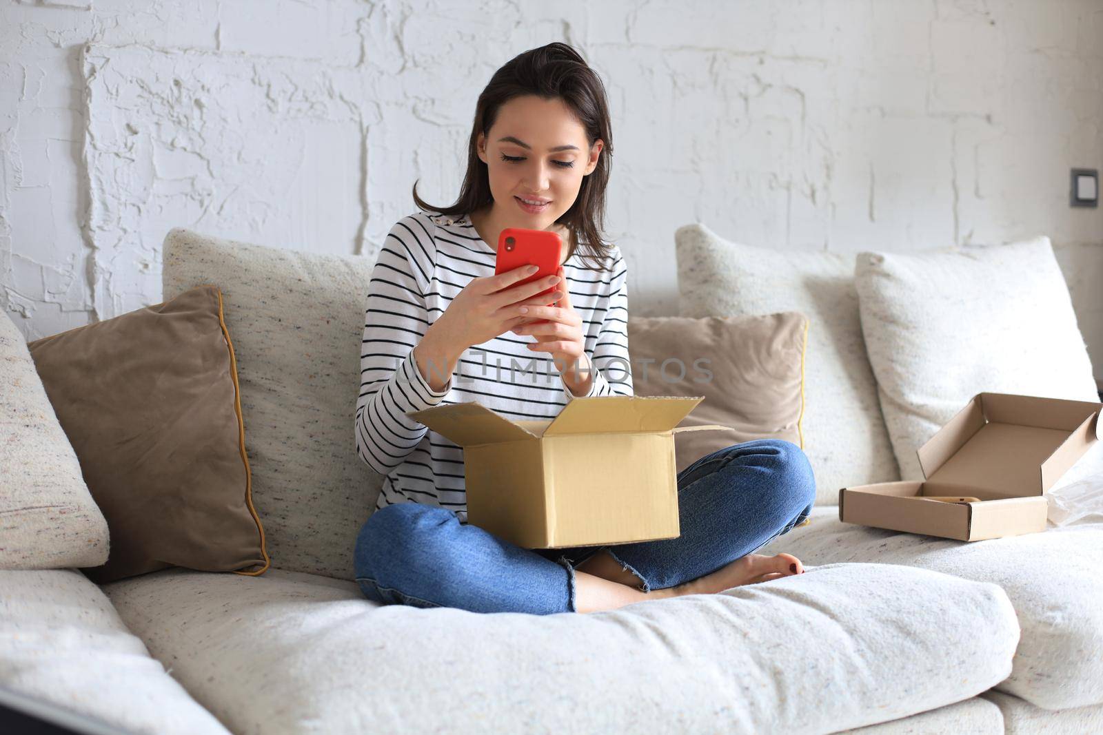 Beautiful young woman is holding cardboard box and unpacking smartphone sitting on sofa at home. by tsyhun