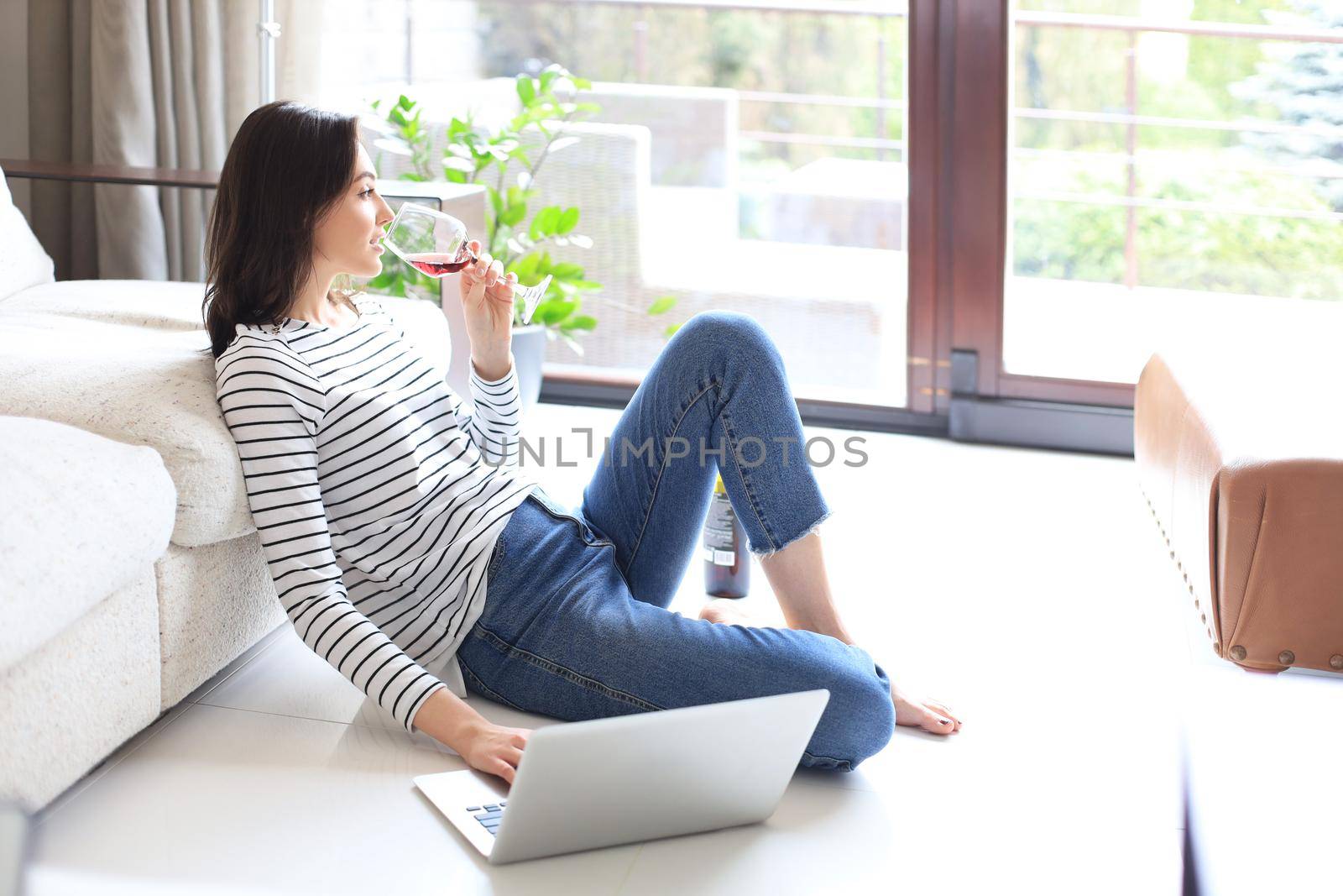 Smiling young woman sitting on floor with laptop computer and chating with friends, drinking wine. by tsyhun