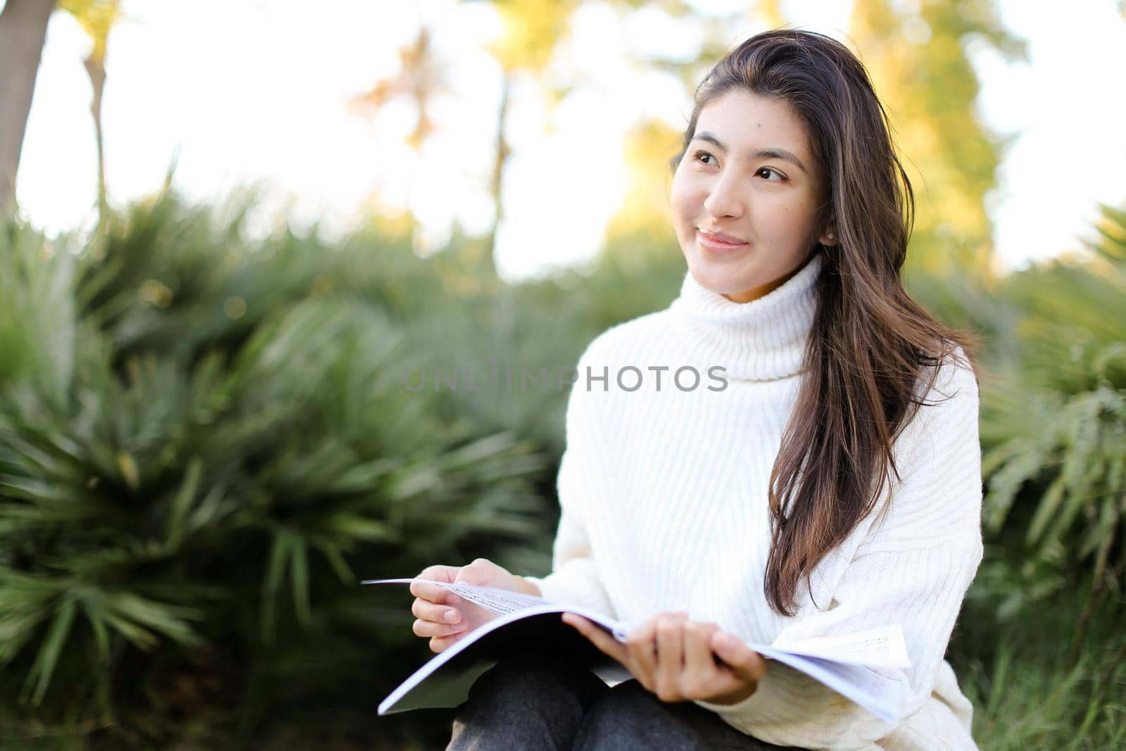 Chinese smiling female student sitting on stump in park and reading papers. Concept of preparing before exam and asian woman.