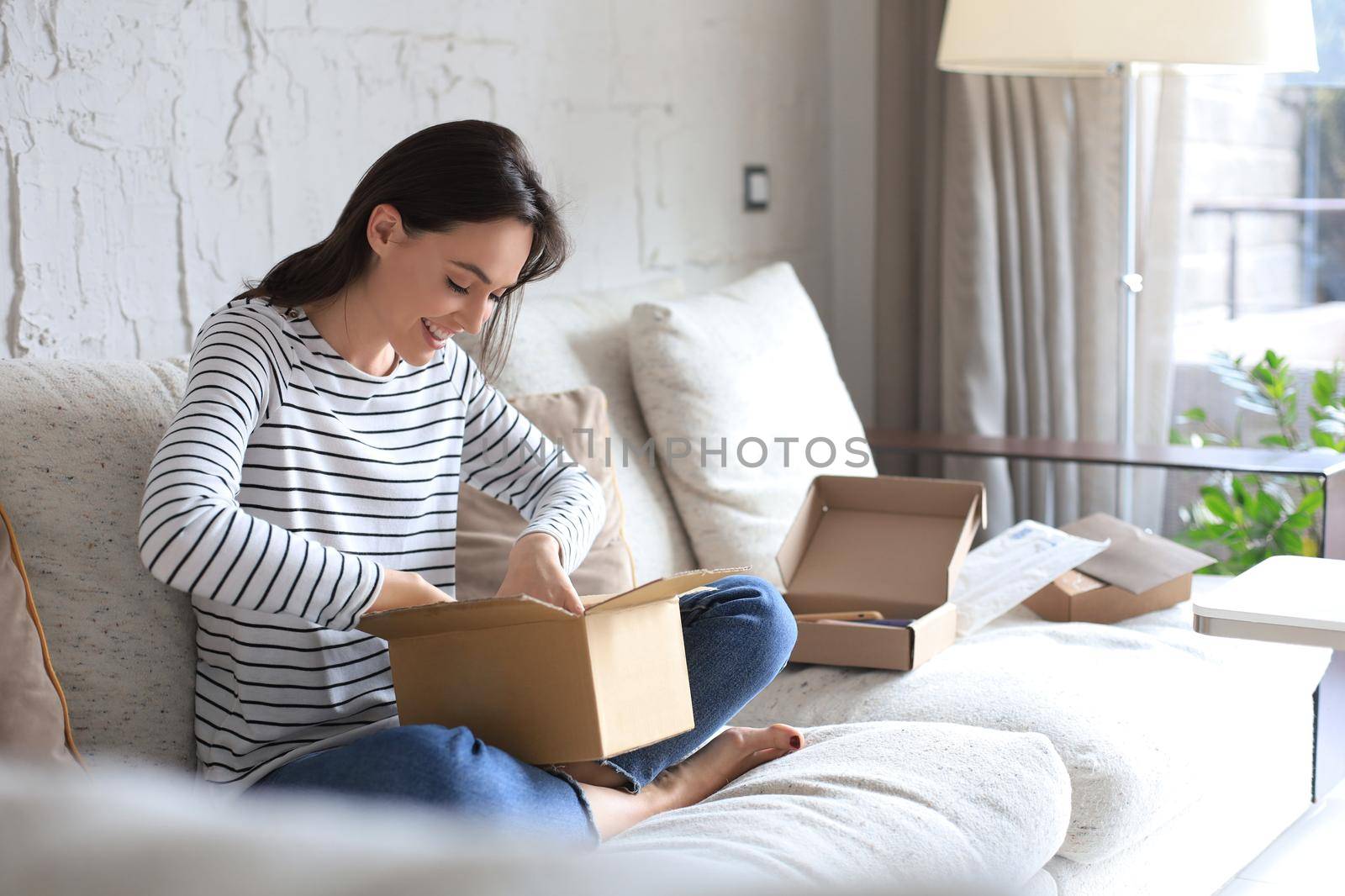 Beautiful young woman is holding cardboard box and unpacking it sitting on sofa at home