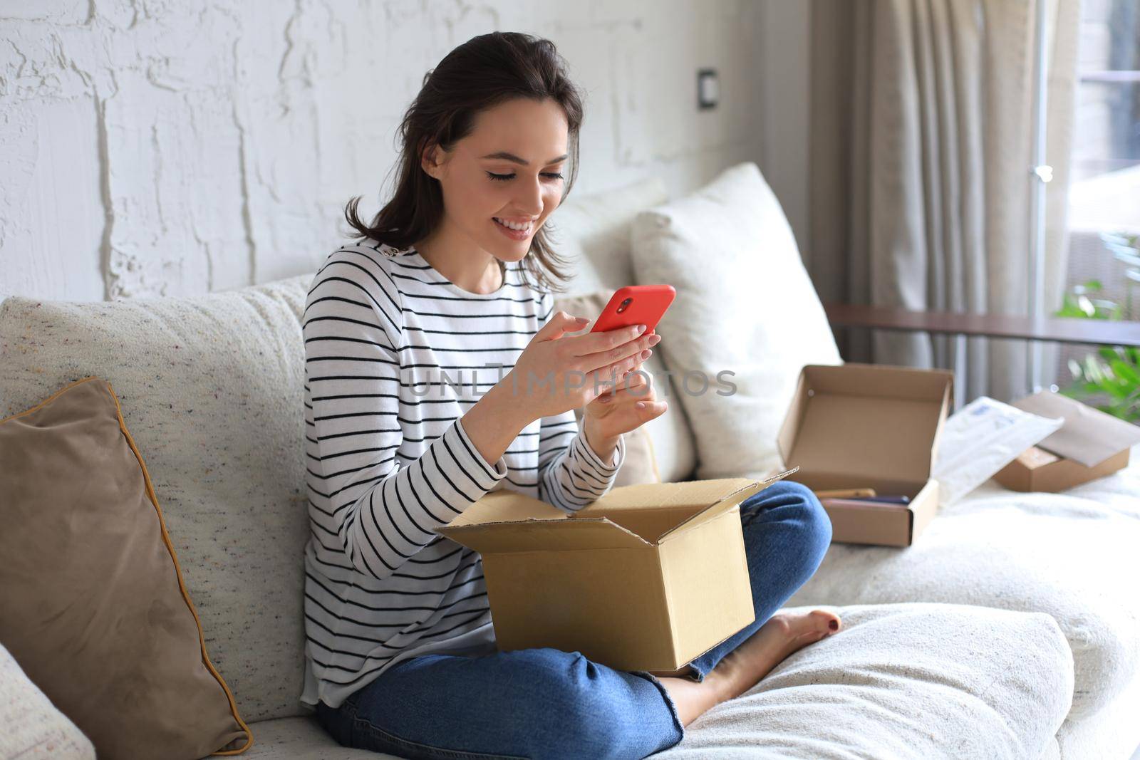 Beautiful young woman is holding cardboard box and unpacking smartphone sitting on sofa at home. by tsyhun