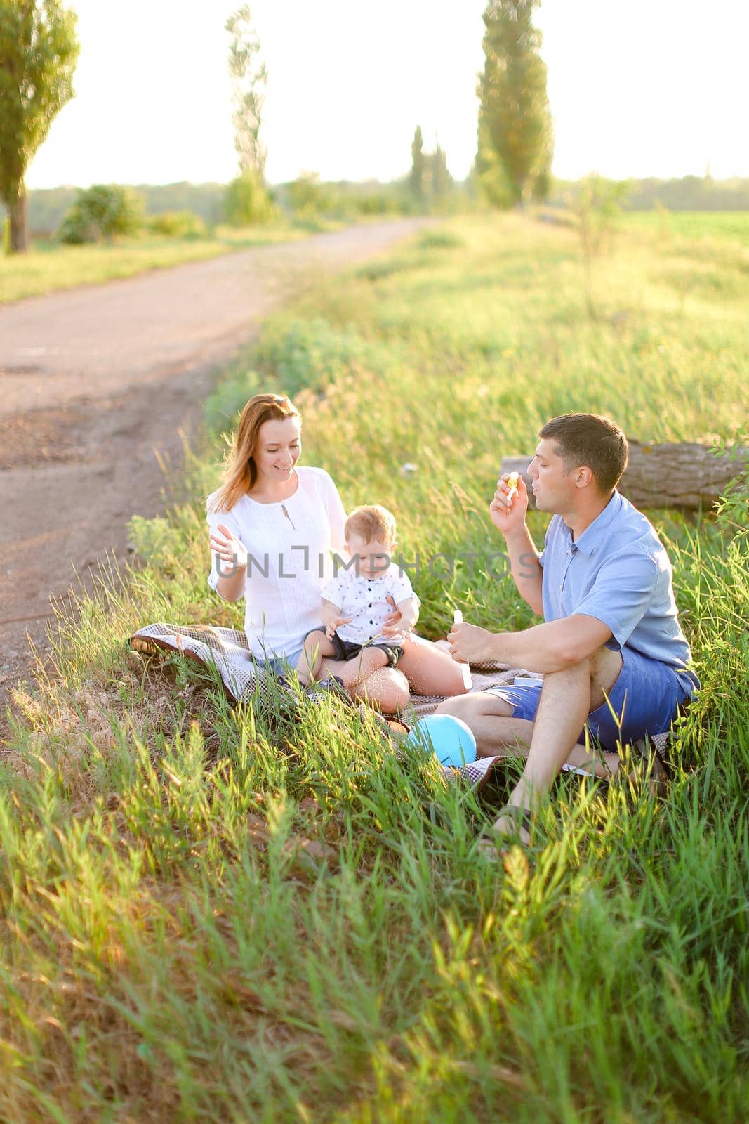 Young father and blonde cute mother sitting on grass with little baby and blowing bubbles. Concept of picnic with child and resting on nature.