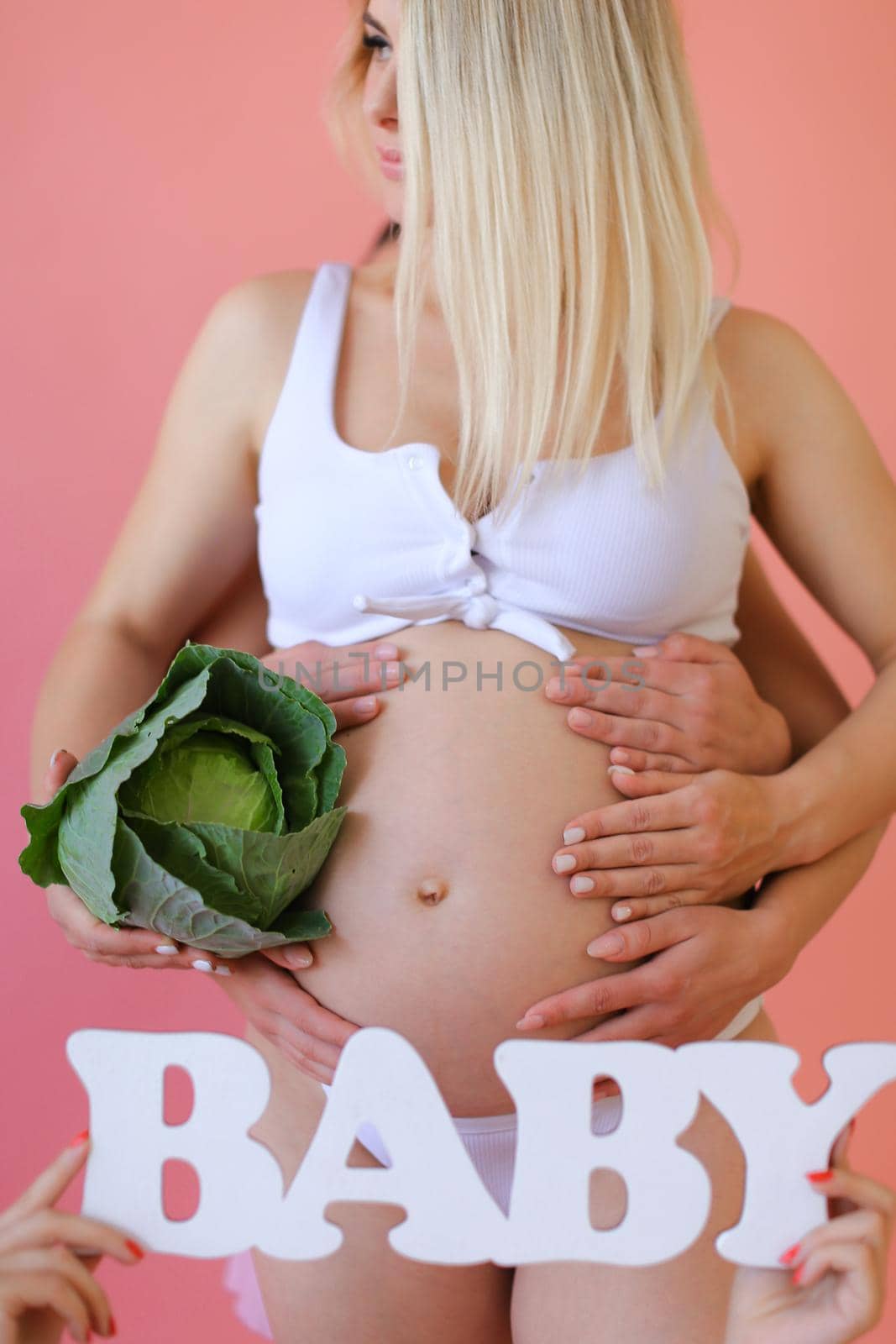 Closeup pregnant blonde woman in underwear keeping inscription baby and cabbage in pink monophonic background. Concept of expactant photo session.