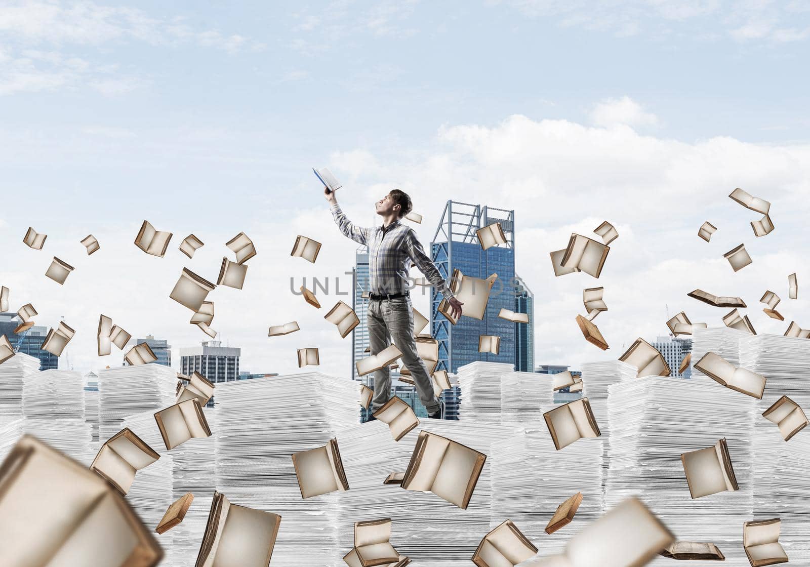 Man in casual wear keeping hand with book up while standing among flying books with cityscape on background. Mixed media.
