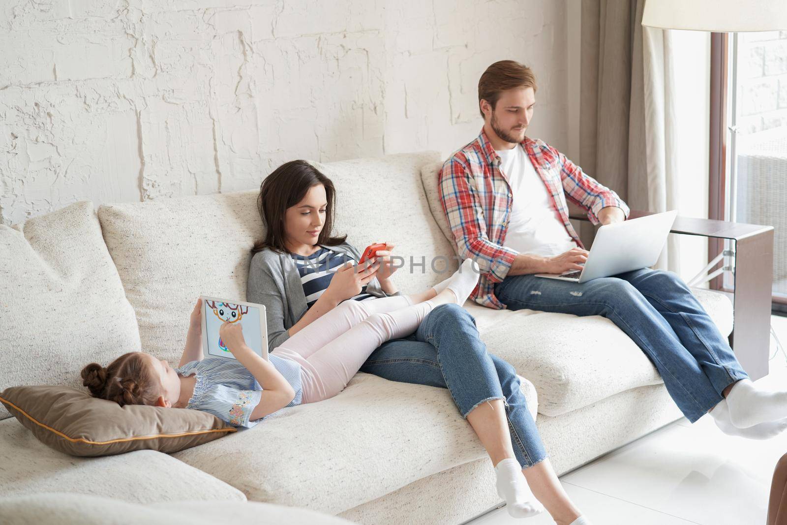 Father, mother and daughter using electronic devices sitting on sofa at living room. by tsyhun