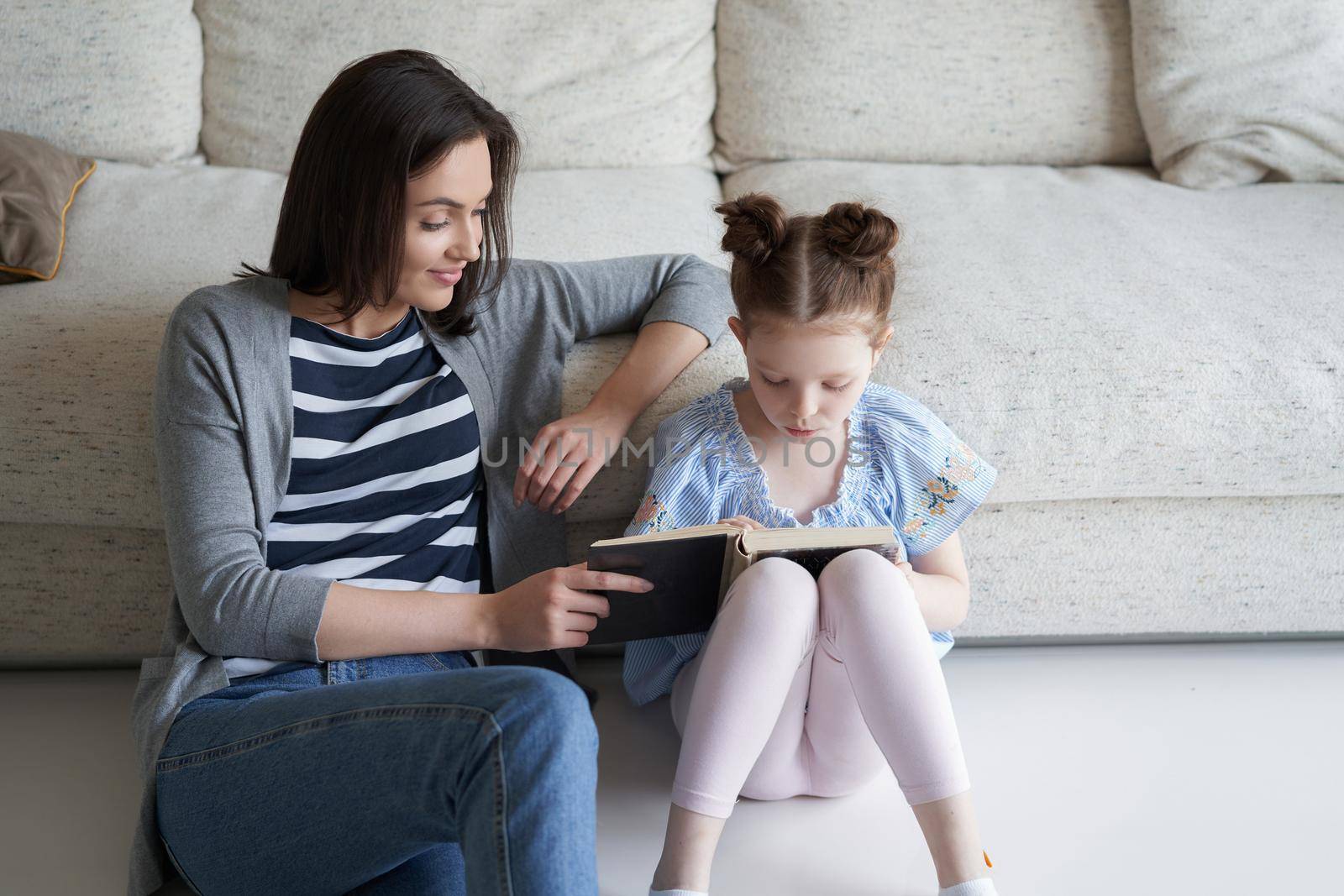 Smiling young mother and little daughter sitting on floor near sofa at home and reading books