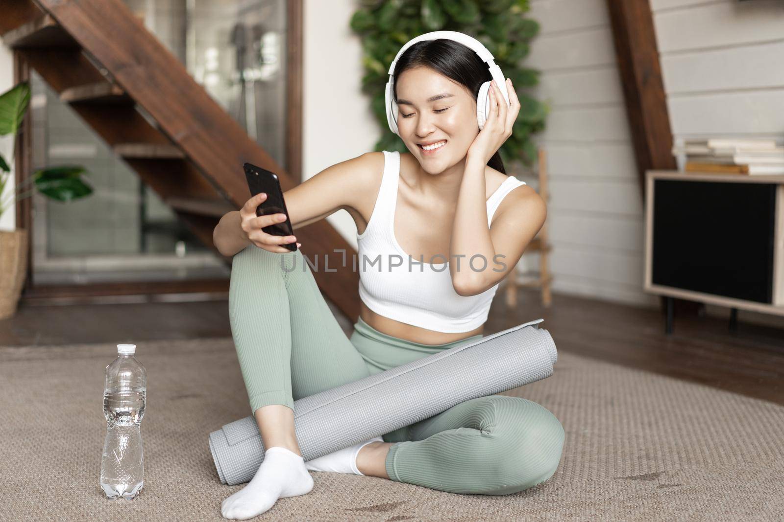 Happy sporty girl sitting on floor at home and taking selfie during workout, wearing headphones and holding smartphone by Benzoix