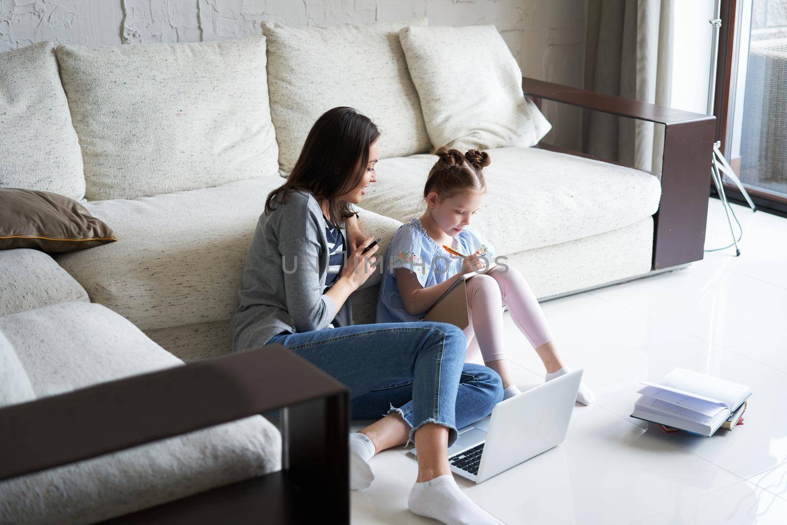 Smiling mother and daughter sitting on floor in living room with laptop, teaching lessons. by tsyhun