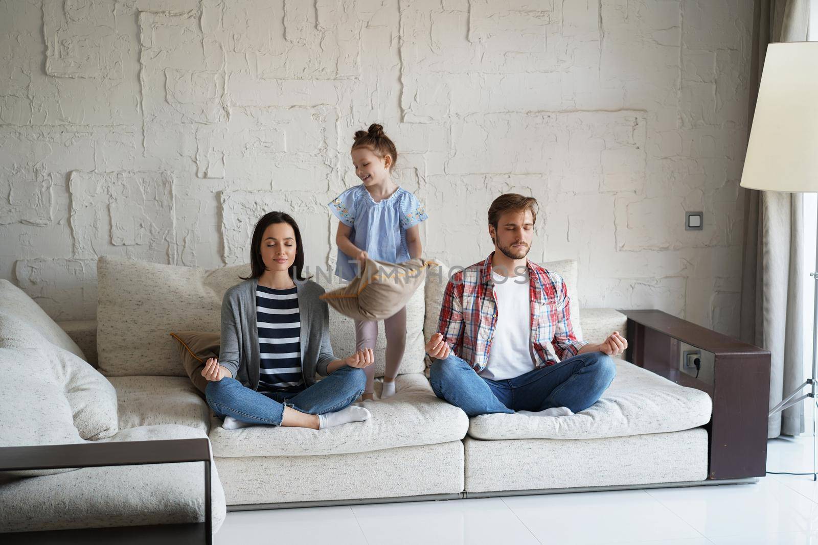 Exhausted mother and father sitting on couch feels annoyed tired while noisy little daughter shouting run around sofa