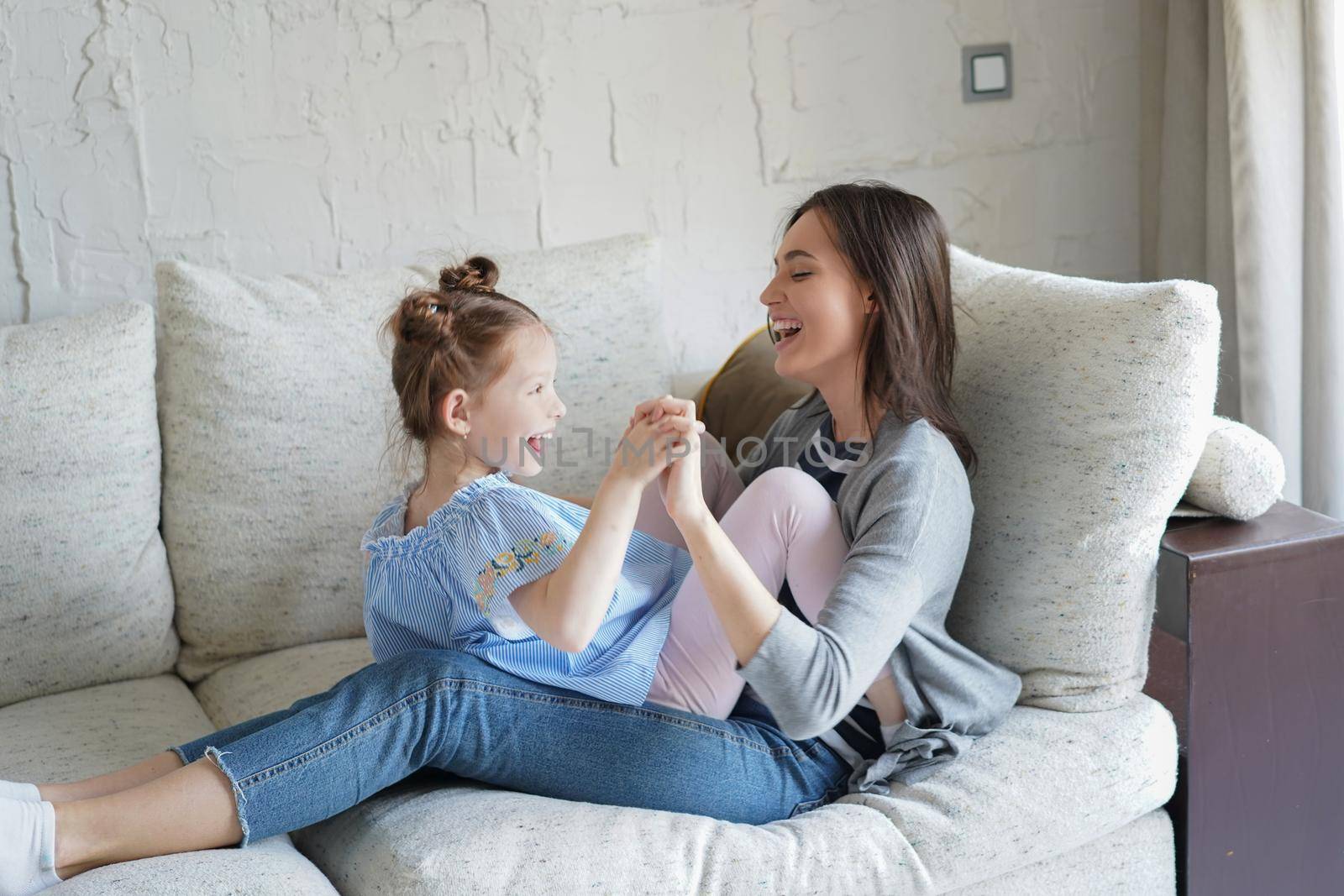 Cute little kid daughter laughing playing with mother on sofa, happy mother relaxing having fun with funny small child girl bonding enjoying leisure together in living room