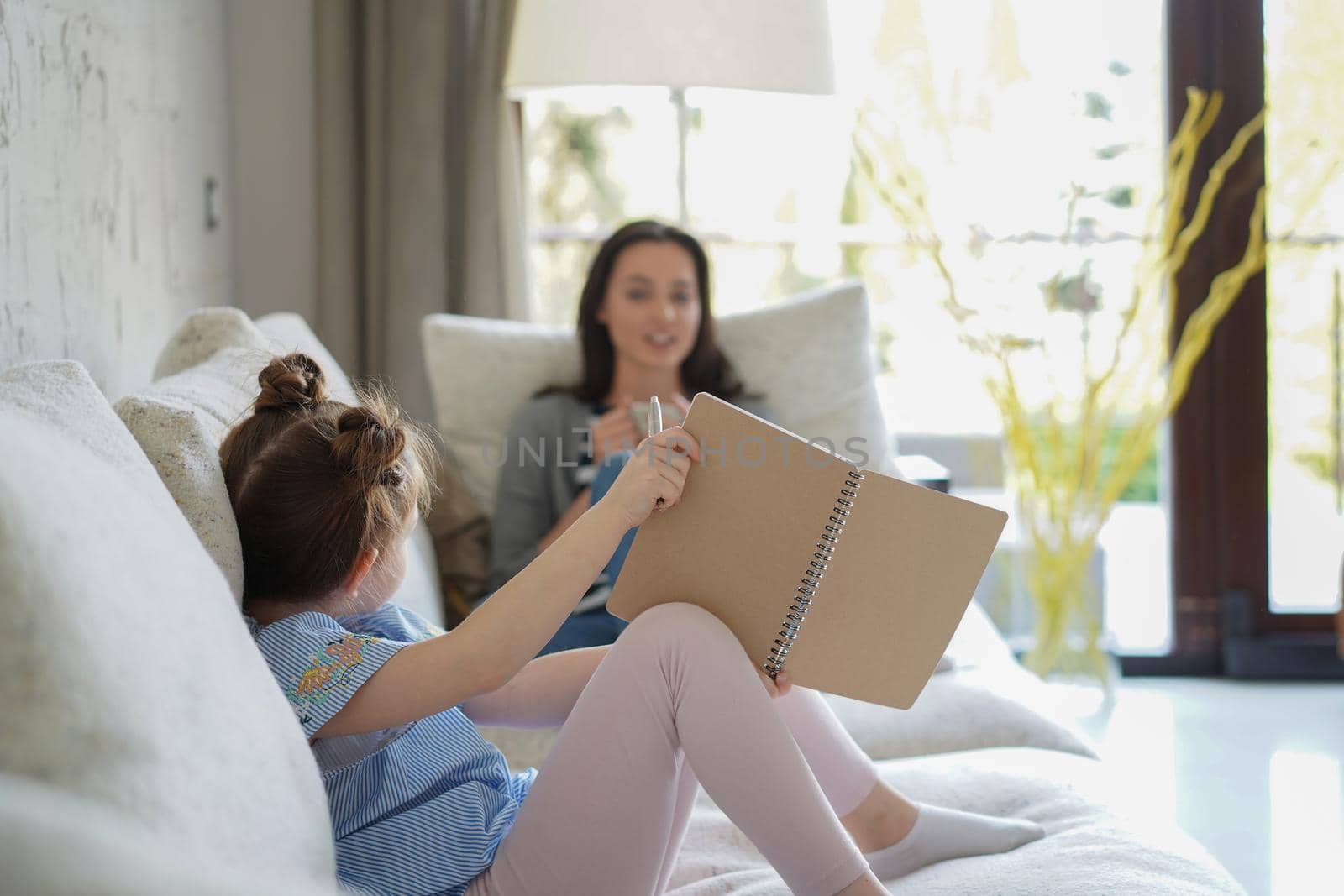 Happy mother smilling daughter sitting on sofa enjoying creative activity, drawing pen pictures in albums, mother and daughter spend free time together. by tsyhun