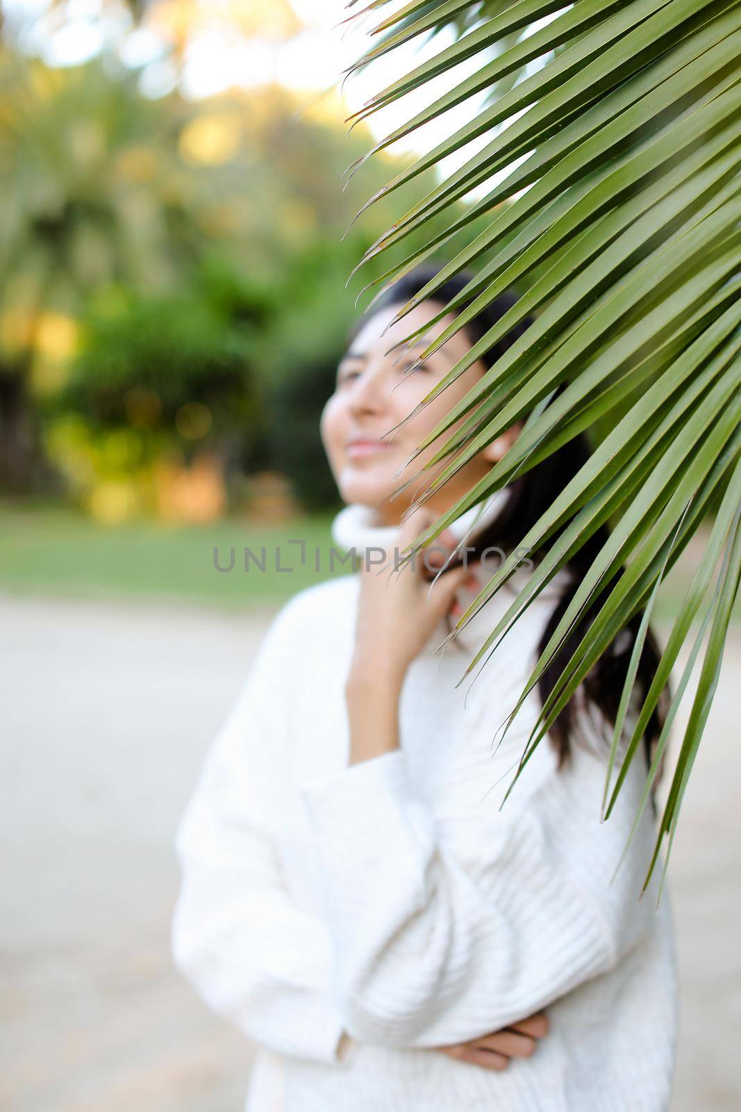 Focus on palm leaf, chinese woman wearing white sweater standing on beach. Concept of asian beauty and tropical nature.
