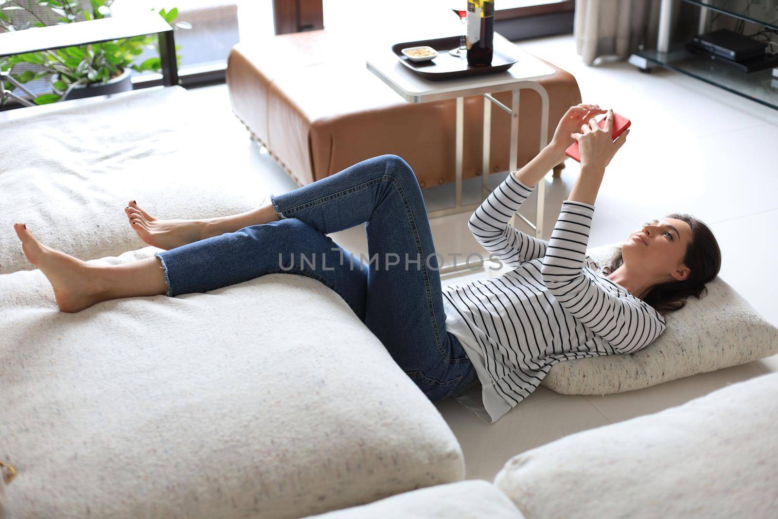 Young pretty woman checking social media, lying on floor against cozy domestic interior, drinking red wine