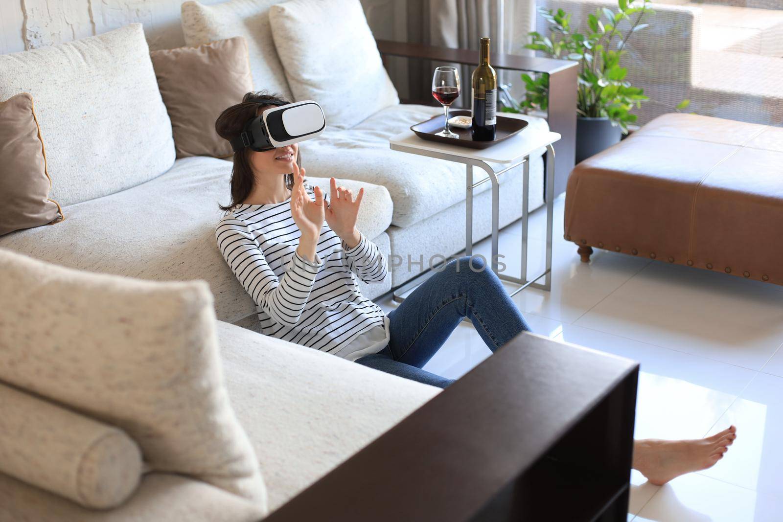 Young casual woman in goggles of virtual reality sitting on floor at home looking around in excitement. by tsyhun