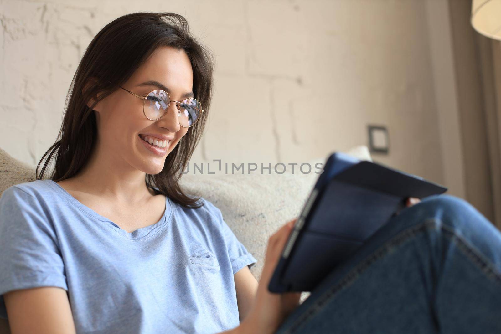 Smiling young woman sitting on sofa with digital tablet and chating with friends