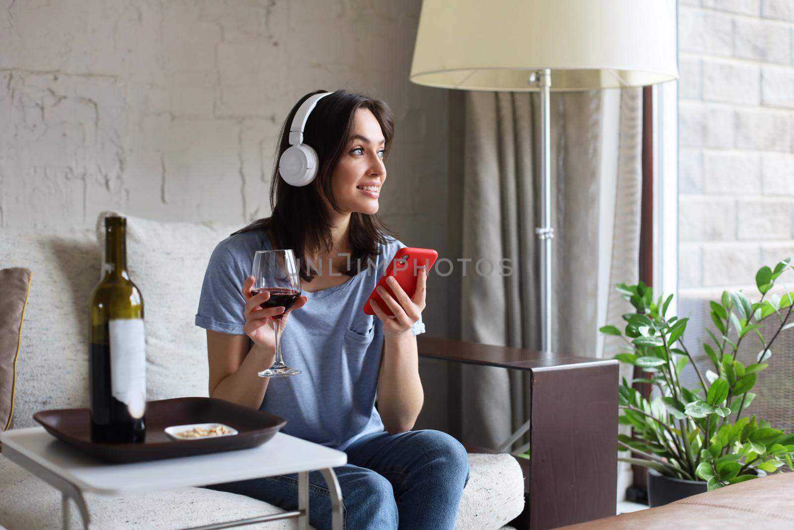 Pretty girl using her smartphone on couch at home in the living room. Listening music, drinking red wine, relaxation after a hard week at work