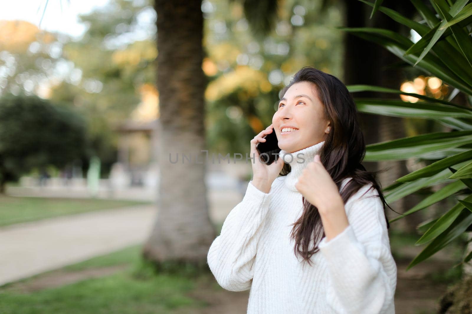 Chinese happy girl in tropical park talking by smartphone near palm. Concept of modern techology and nature.