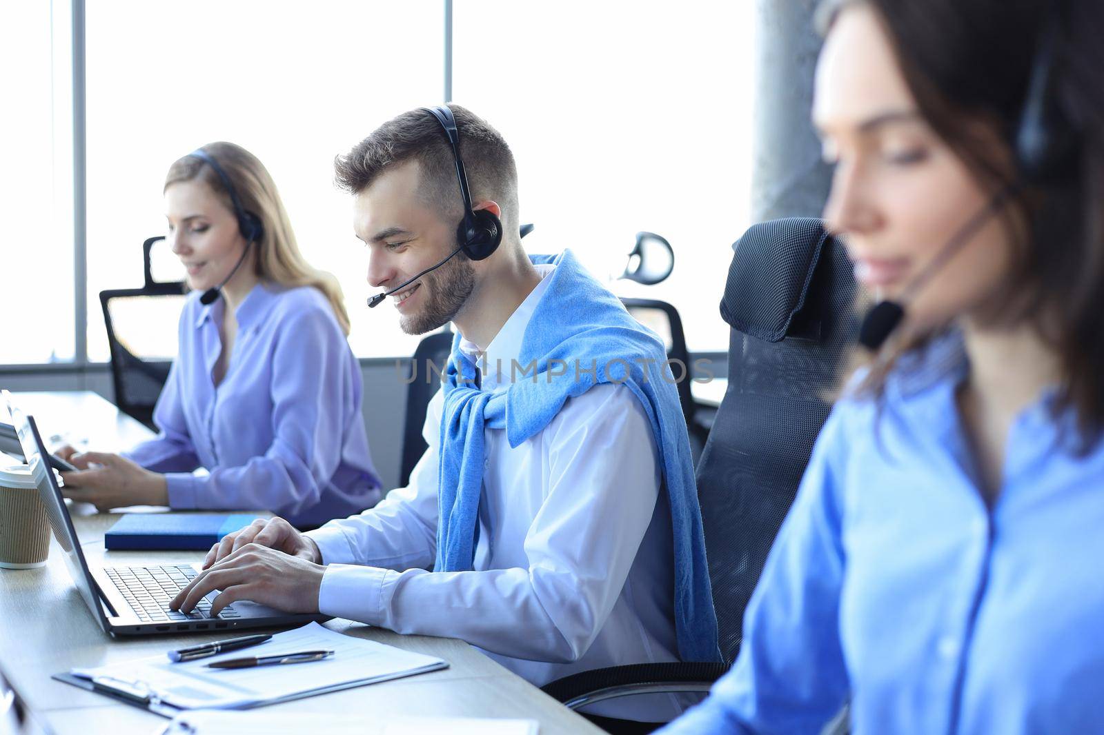 Smiling male call-center operator with headphones sitting at modern office with collegues on the backgroung, consulting online