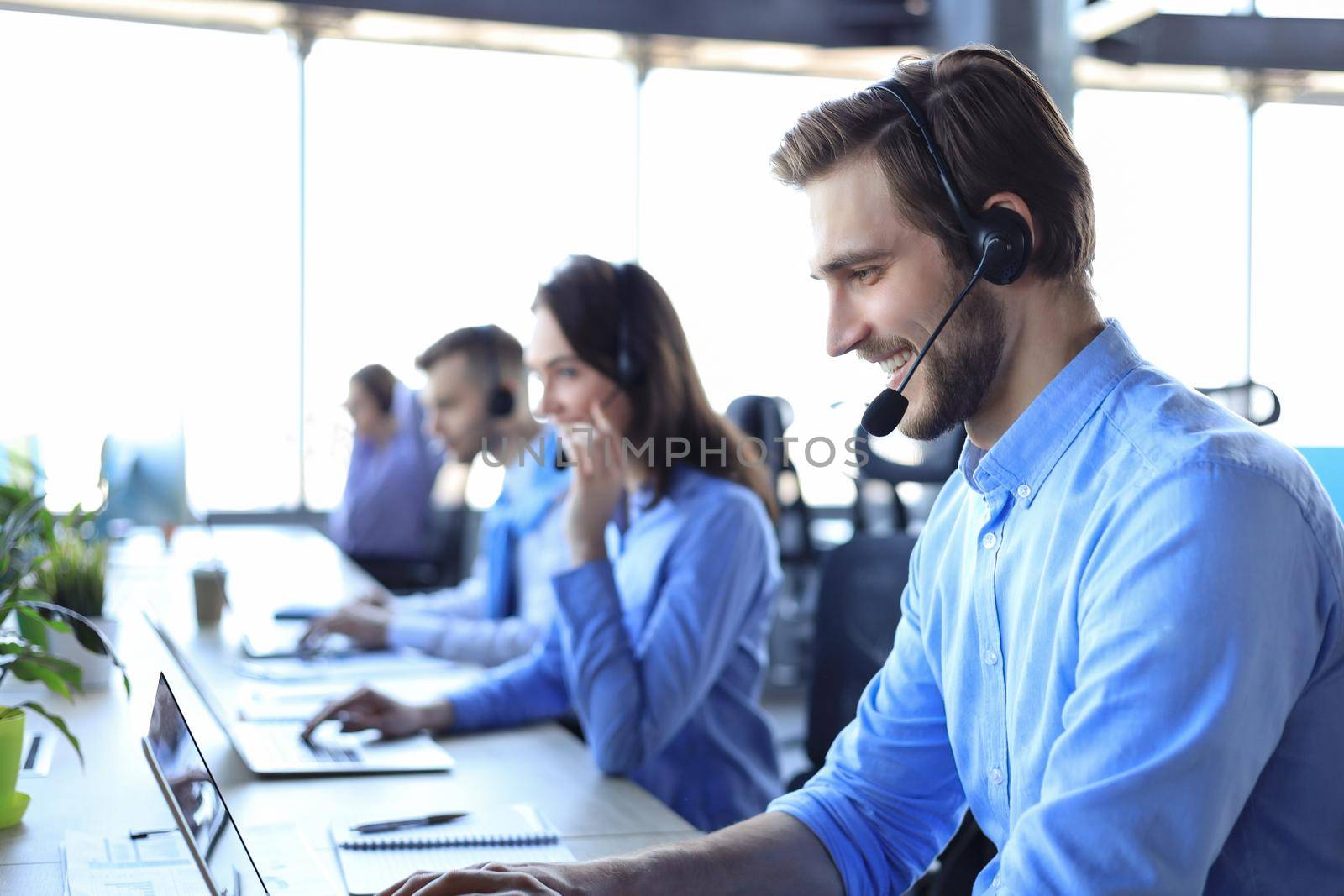 Smiling male call-center operator with headphones sitting at modern office with collegues on the backgroung, consulting online. by tsyhun