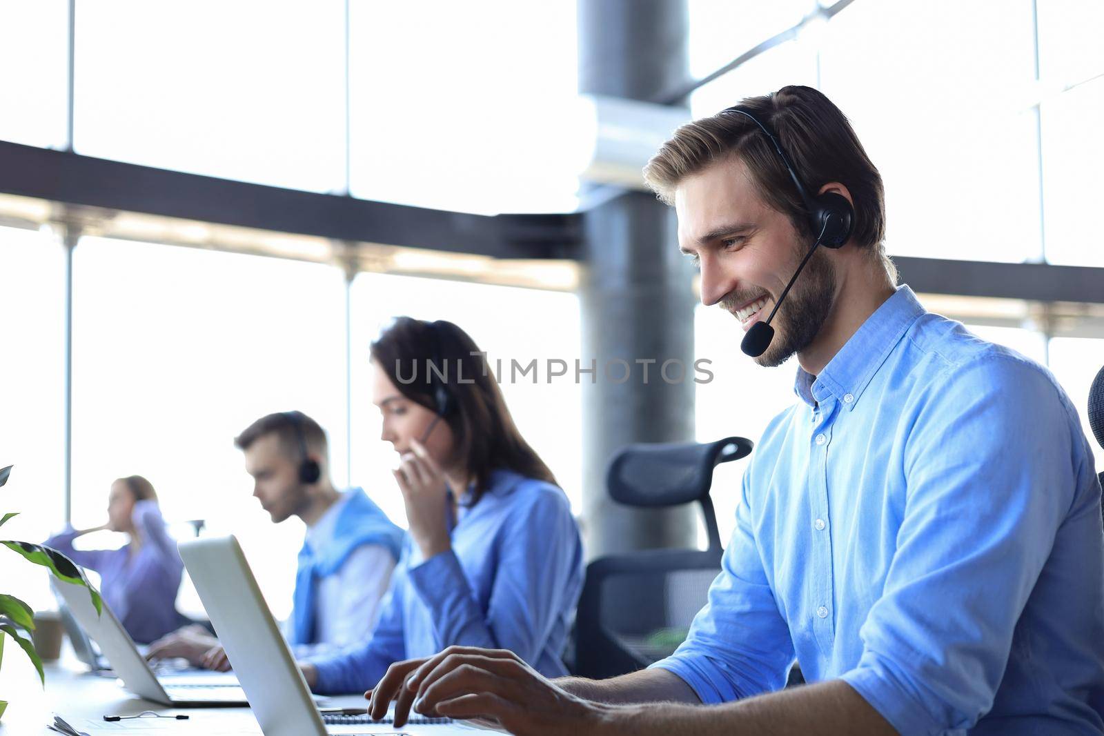 Smiling male call-center operator with headphones sitting at modern office with collegues on the backgroung, consulting online. by tsyhun