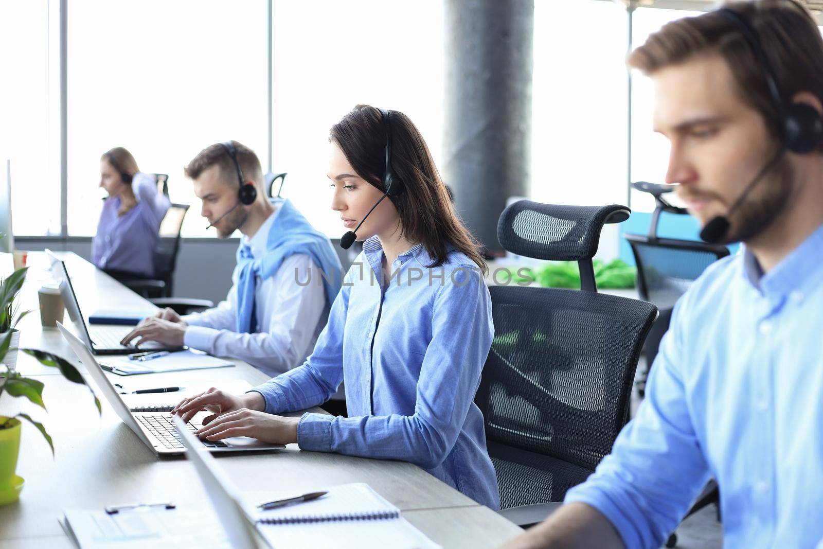 Smiling male call-center operator with headphones sitting at modern office with collegues on the backgroung, consulting online. by tsyhun