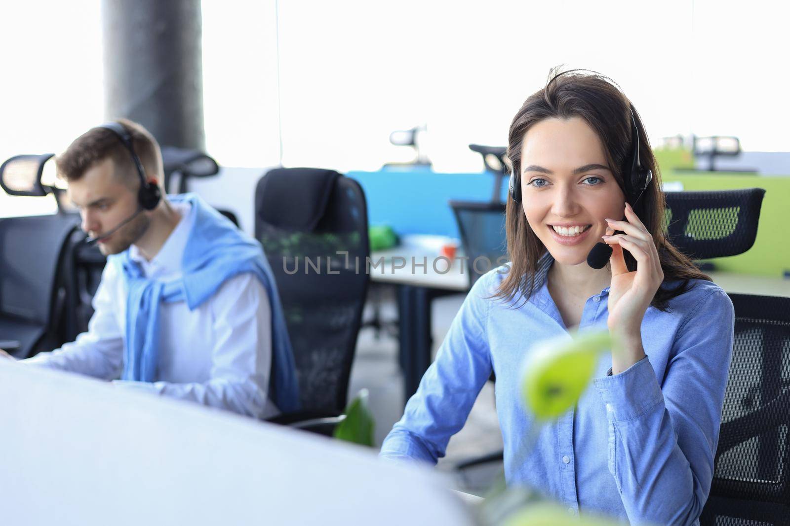 Smiling male call-center operator with headphones sitting at modern office with collegues on the backgroung, consulting online. by tsyhun