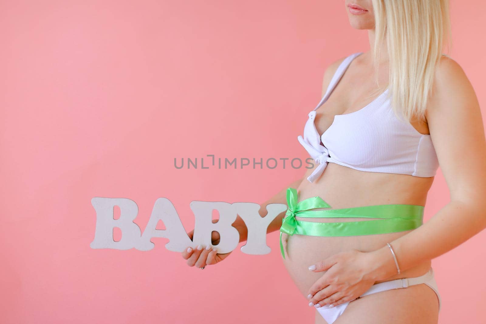 Closeup pregnant female person in underwear with ribbon on belly keeping inscription baby in pink monophonic background. Concept of expactant photo session.