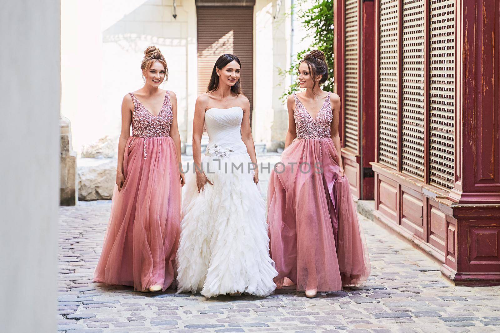 Beautiful bride and bridesmaids walking on the cobblestone street. Maids wear gorgeous elegant stylish red pink violet floor length v neck chiffon gown dress decorated with sequins sparkles and rhinestones. Wedding day in old beautiful European city. by berezko