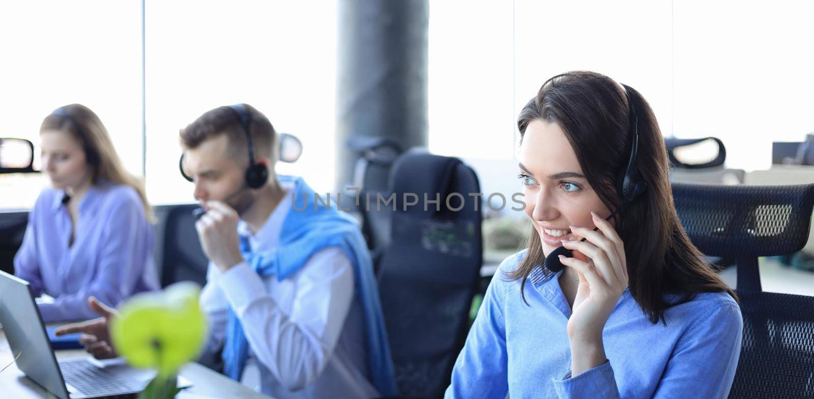 Female customer support operator with headset and smiling, with collegues at background. by tsyhun