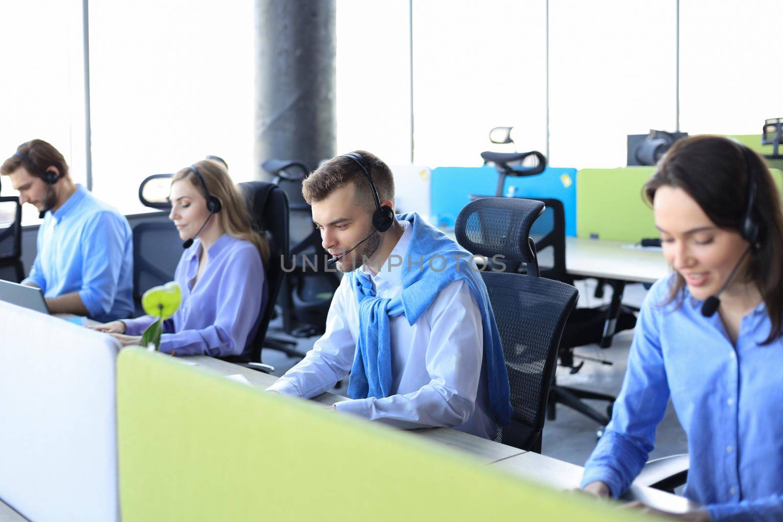 Smiling male call-center operator with headphones sitting at modern office with collegues on the backgroung, consulting online