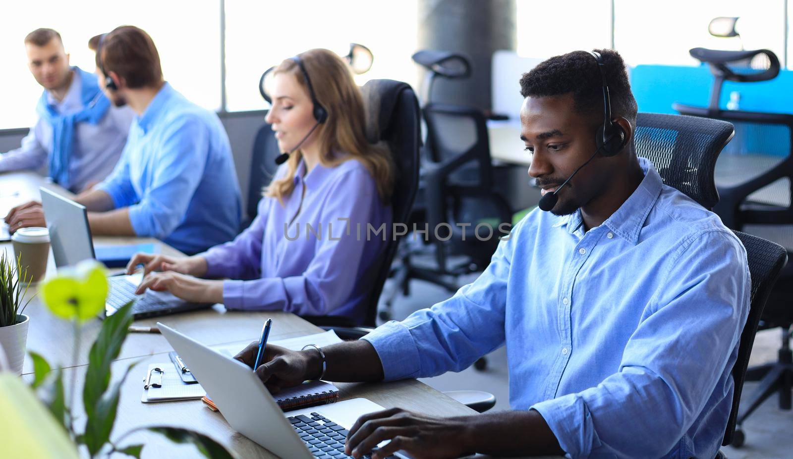 Smiling african male call-center operator with headphones sitting at modern office with collegues on the backgroung, consulting online