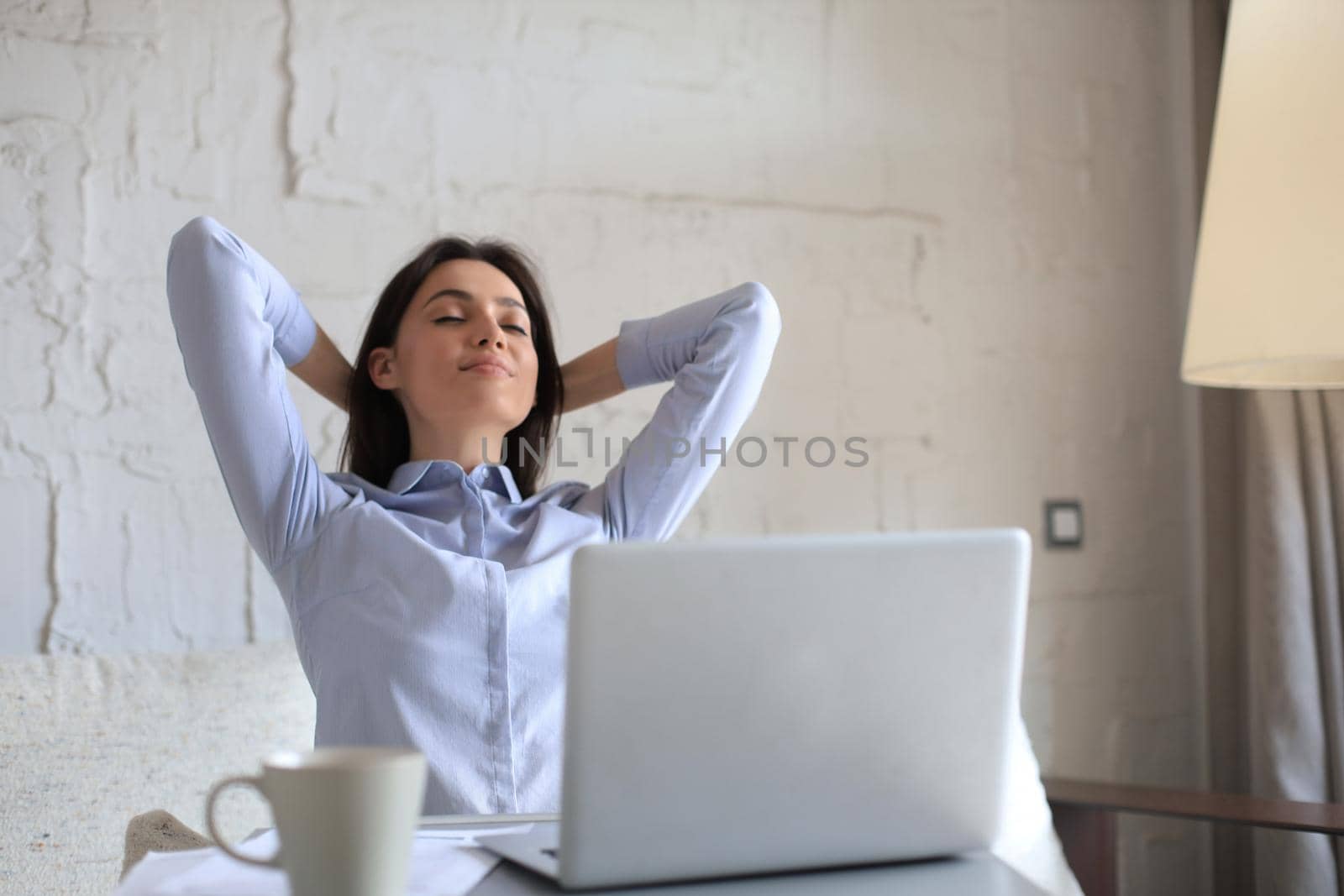 Young woman crossed hands behind head, enjoying break time at home. Peaceful carefree business woman resting at table with computer, looking aside, dreaming of future