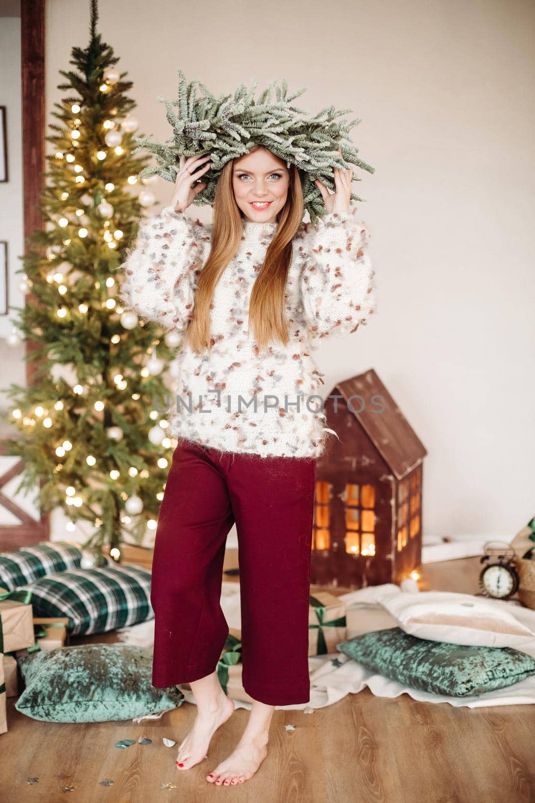 Full-length photo of a red-haired woman with a crown of fir branches posing near Christmas tree