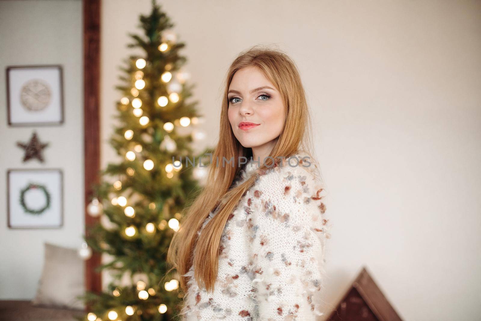 Waist-up photo of a stunning red-haired lady standing near Christmas tree and looking at camera