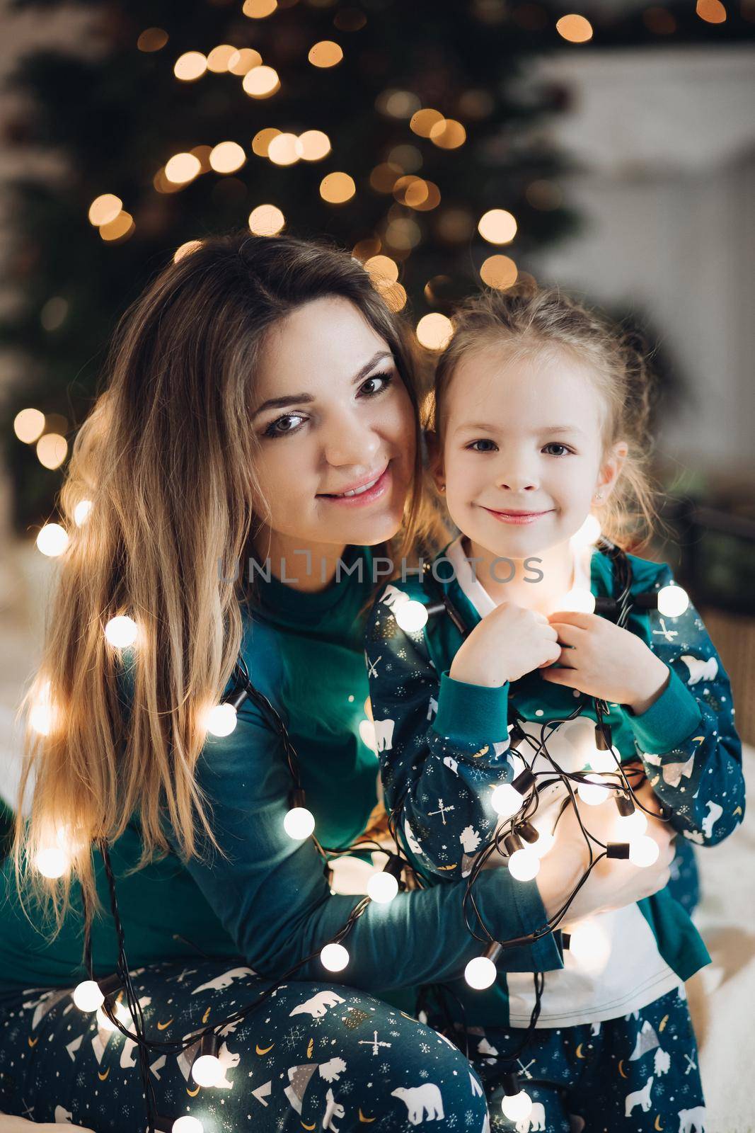 Beautiful young lady and lovely little girl with shiny Christmas lights around their shoulders posing for a camera. Holiday concept
