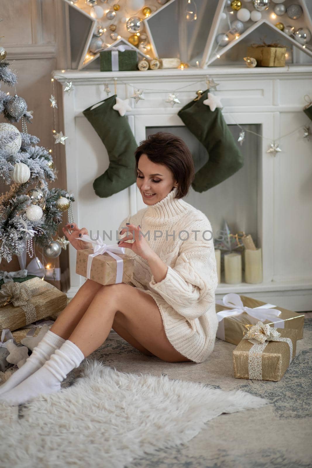 Portrait of a pretty adult woman in white knitted sweater opening Christmas gift sitting on floor.