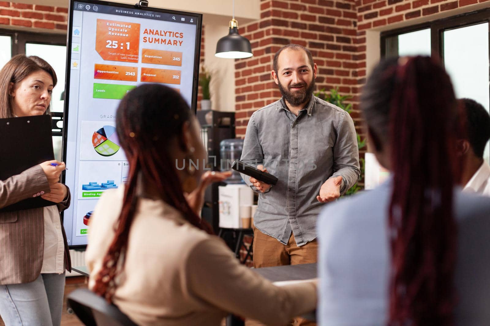 Executive manager standing beside monitor showing marketing graphs explaining company strategy during business meeting in startup office. Diverse businesspeople working at financial presentation