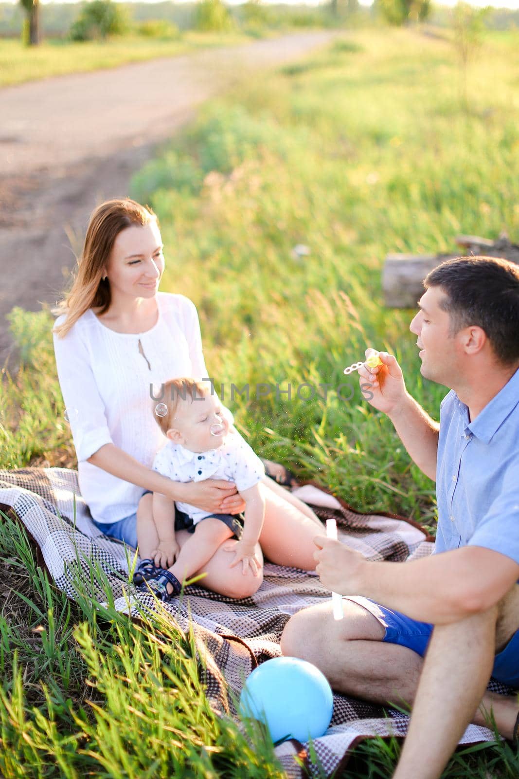 Young happymother and father sittling on grass with little baby and blowing bubbles. Concept of picnic and children, parenthood and leisure time.