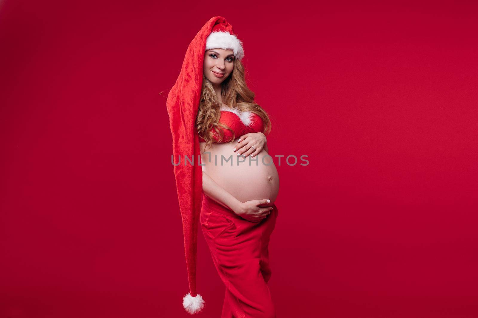 Young beautiful pregnant woman in Santa costume hugs her stomach by StudioLucky