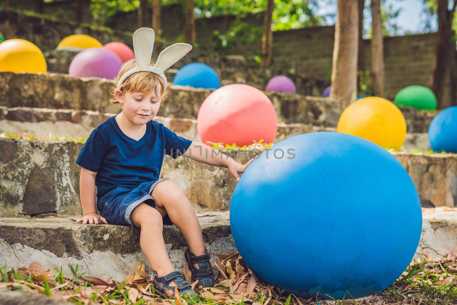 Cute little kid boy with bunny ears having fun with traditional Easter eggs hunt, outdoors. Celebrating Easter holiday. Toddler finding, colorful eggs by galitskaya