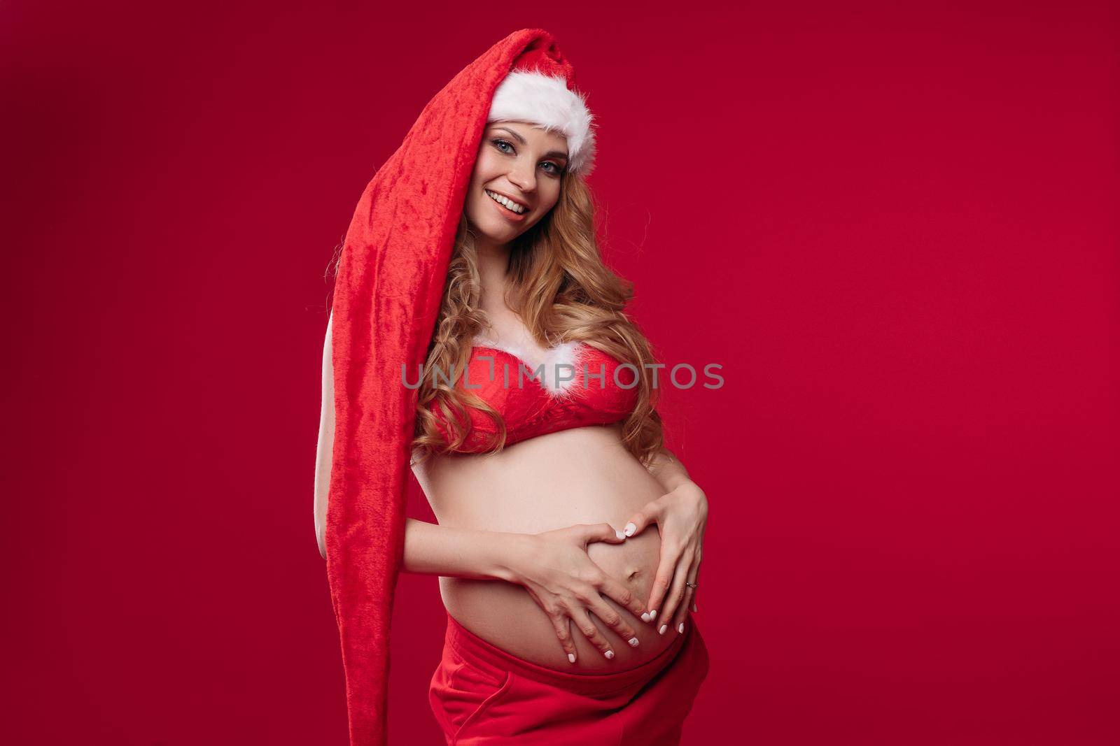 Charmilng young pregnant female in christmas costume smiles, picture isolated on red background