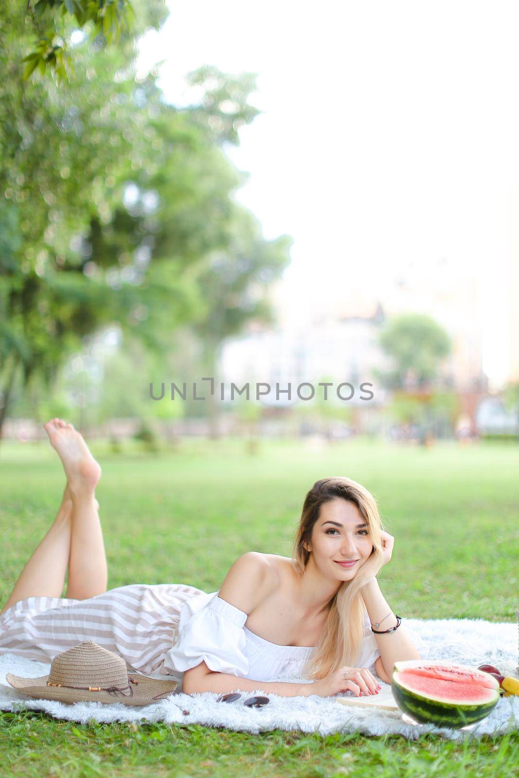 Young blonde eueropan woman reading book and lying on plaid near fruits and hat, grass in background. Concept of summer picnic and resting on weekends in open air.