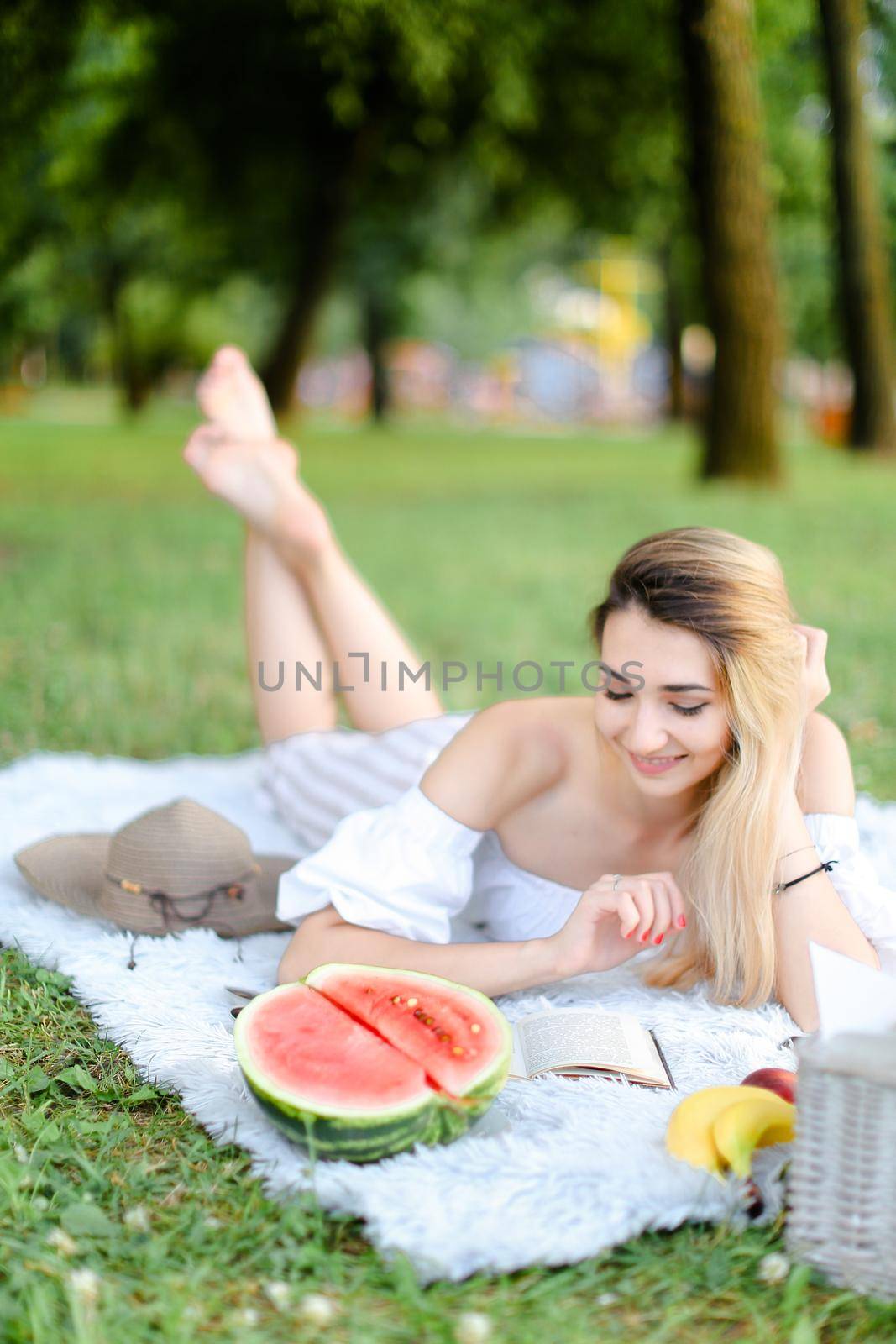 Young blonde pretty woman lying on plaid in park, reading book near watermelon and hat. Concept of leisure time, summer vacations and picnic.