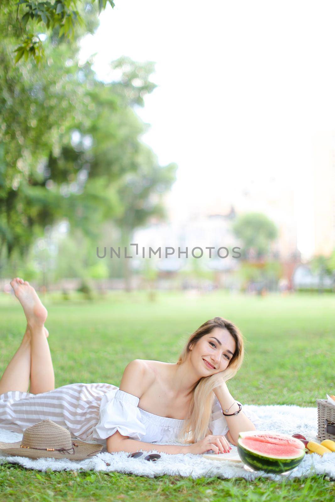 Young blonde woman reading book and lying on plaid near watermelon and hat, grass in background. Concept of summer picnic and resting on weekends in open air.